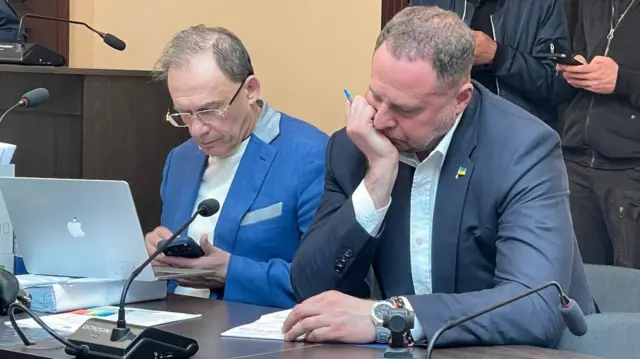 За столом у суді сидять дві людини. Ліворуч – адвокат Фомін, перед ним – відкритий макбук, у руках – мобільний телефон. Праворуч – Єрмак, однією рукою підпирає голову; на столі перед ним – аркуш із записами й мікрофон. На задньому плані стоять ще кілька людей у темному одязі.