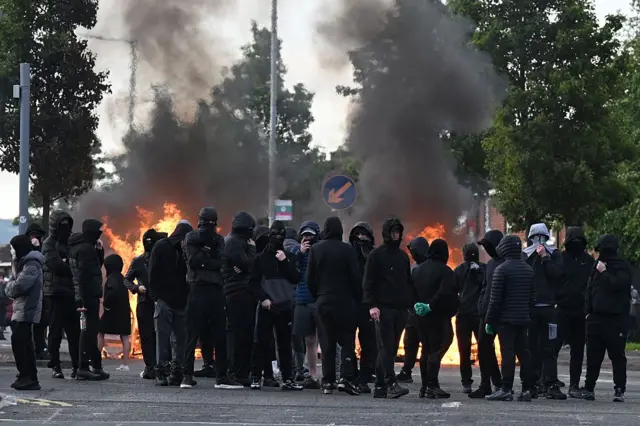 Jóvenes encapuchados con fuego detrás.