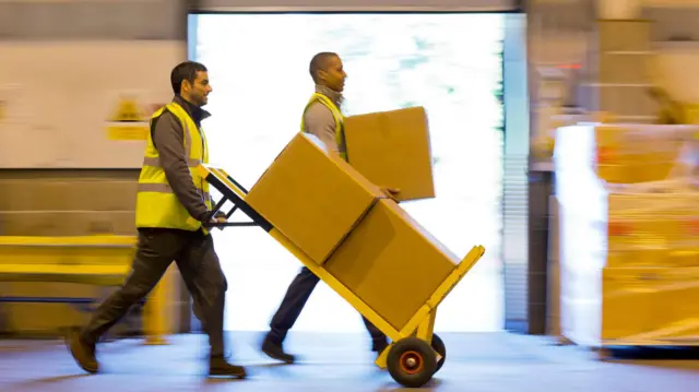 黄色いジャケットを着た男性2人が倉庫の中を歩いている。1人は茶色の段ボール箱を両手で持ち、もう1人は同じ大きさの段ボール2個を載せた台車を押している