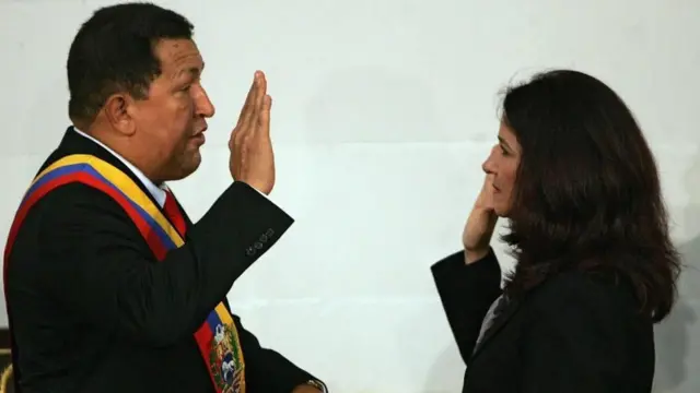 Hugo Chavez holds up his right hand facing Flores, who does the same in what appears to be a swearing-in ceremony. Both wear dark suits and have serious expressions.