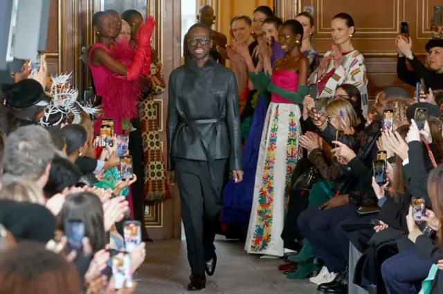 A man walks down a catwalk dressed in black and smiles at seated spectators. Behind him are models dressed in this clothing.