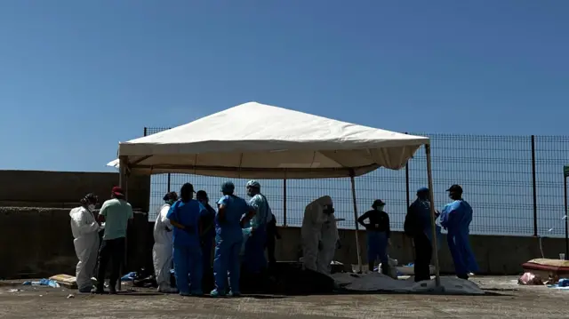 Personal médico alrededor de cadáveres sobre el suelo cubiertos con bolsas de plástico. Los trabajadores llevan batas azules o blancas. 