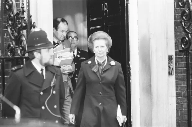 Margaret Thatcher saliendo de Downing Street, con dos funcionarios detrás de ella y un policía delante.