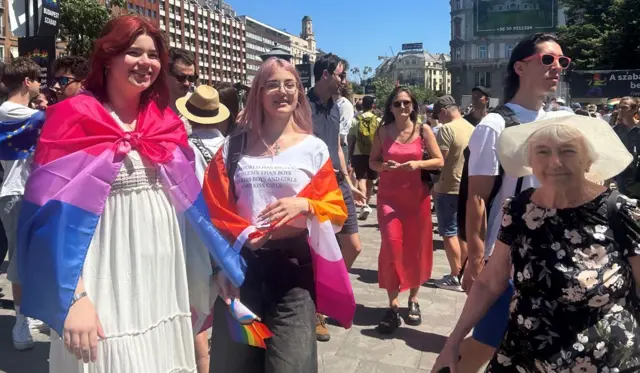 ブダペストの街中で、カラフルな布地を肩に巻いた若い女性たちの隣に、花柄のワンピースと白い麦わら帽子をかぶった高齢の女性が立っている