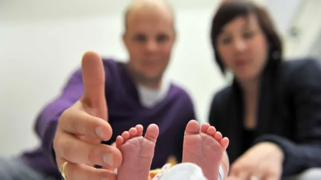 Muškarac i žena gledaju u bebu, muškarac prstima dotiče bebina stopala koja se jedina vide na ekranu