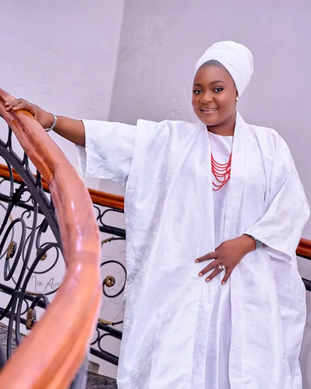 aworan yii ni aworan Bunmi Akinnaanu ninu aṣọ funfun pẹlu gele funfun, o duro lẹnu akasọ inu ile kan o si fi ọwọ otun di akasọ naa mu.