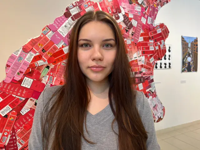 Una mujer joven, de pelo largo y castaño y un jersey gris con cuello en V, se encuentra frente a una sección de la escultura cubierta de paquetes de vapeo rojos y aplanados.