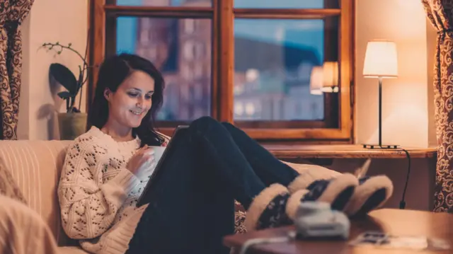 Kobieta o brązowych włosach, ubrana w biały sweter, dżinsy i puszyste buty, siedzi na sofie i patrzy na swój tablet.