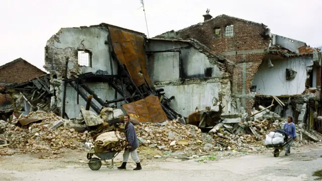 Ethnic Albanians push wheelbarrows wey contain dia property in front of a ruined building, April 1999