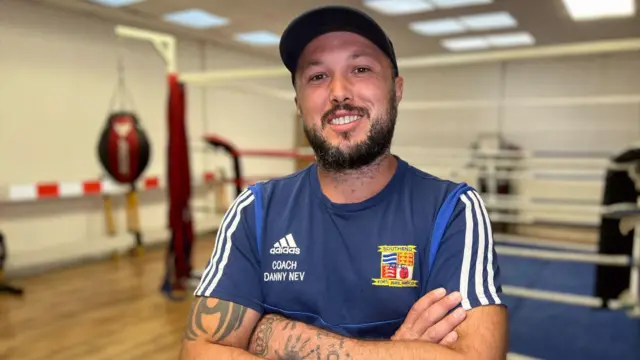 Danny Neville lleva una camiseta azul marino con el logo de Adidas en el pecho derecho y la inscripción "Coach Danny Nev" debajo. En el pecho izquierdo luce el escudo del Southend Amateur Boxing Club. Lleva una gorra de béisbol oscura y barba oscura, y está de pie frente a un ring de boxeo.