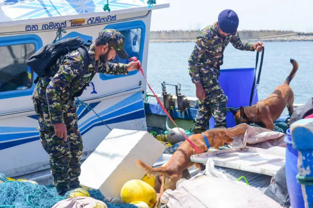 මුහුදේ ඇති බහුදින ධීවර යාත්‍රාවක් නාවික හමුදා නිලධාරීන් දෙදෙනෙකු සුනඛයින් දෙදෙනෙකු යොදා ගෙන සෝදිසි කරන අයුරු