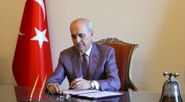 Mavi takım elbiseli bir erkeki, kırmızı türk bayrağının yanında deri bir koltukta oturuyor. Önündeki kağıda imza atıyor.