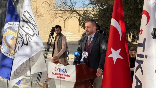 سفیر ترکیه و یک خبرنگار با کامره در این تصویر دیده می شود و سفیر ترکیه کنار پرچم ترکیه ایستاده است 