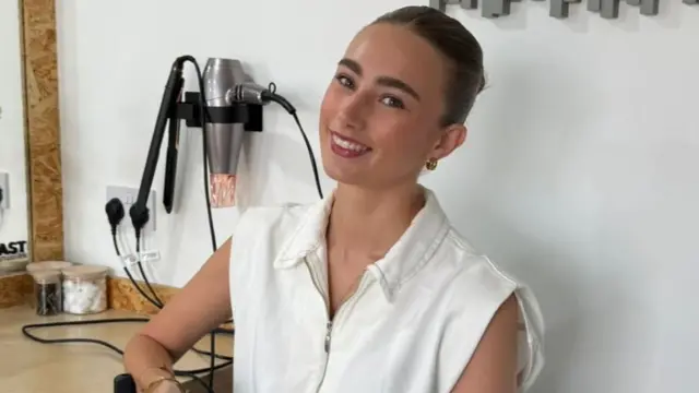 Жінка в білому топі з зав'язаним волоссям посміхається в камеру. Позаду неї — випрямляч для волосся та фен.