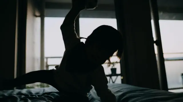 Silhouette image of child crawling across bed wit one arm raised as if to hit out. Di rest of di room dey blurred for di background.