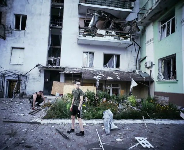 Хлопець, на вигляд приблизно 12 років, стоїть на тлі постраждалої багатоповерхівки: під ногами в нього уламки дерева та металу, поруч лежить зігнутий шматок металу. На перших двох поверхах будинку вибиті всі вікна, на даху крильця лежить вибита віконна рама. Біля будинка чоловік пиляє деревʼяні балки