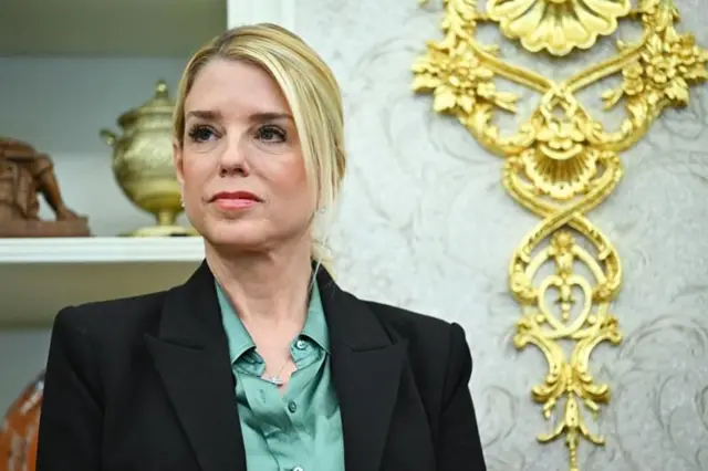Pam Bondi olhando para a frente. Ela é uma mulher loira de meia idade e veste um terninho preto, com camisa verde por baixo