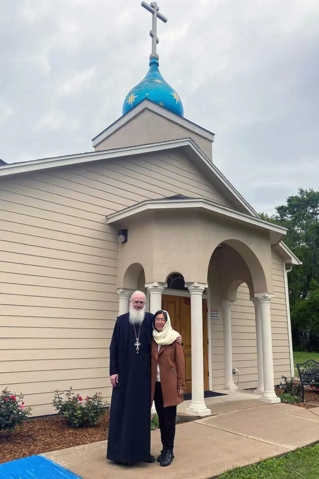 El arcipreste John Whiteford, fotografiado con su esposa Patricia, se encuentra frente a la iglesia ortodoxa de San Jonás en Spring, Texas. Luce una larga barba blanca, viste túnicas negras y lleva una gran cruz alrededor del cuello.