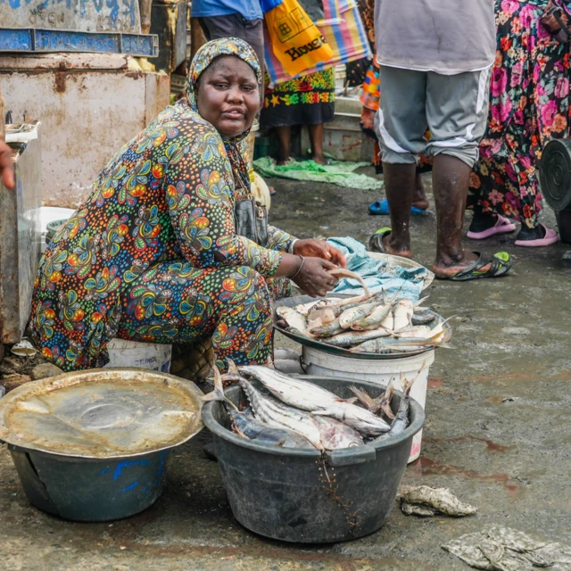 Une femme vêtue d'habits colorés assise près de seaux remplis de poissons sur un marché.

