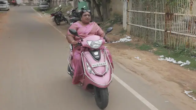 تصویر جایاچترا با ساری صورتی که موترسایکل برقی صورتی سوار شده است. 