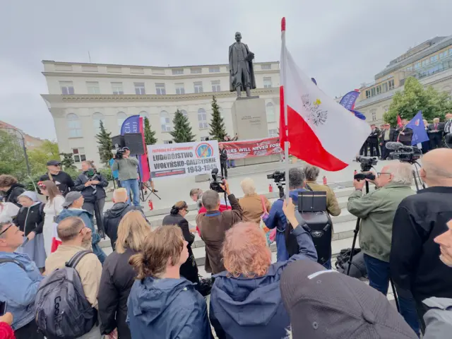 На міській площі біля пам’ятника зібрався натовп людей. У центрі видно сцену з промовцями, журналістами та телекамерами. Над площею майорить великий польський прапор, а біля пам’ятника розгорнуті банери, присвячені пам’яті жертв Волинської трагедії.