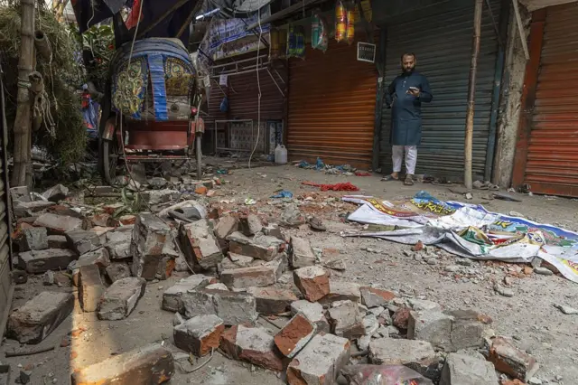 ভূমিকম্পের পর ঢাকার একটি রাস্তায় ছড়িয়ে আছে ভাঙা ইট, সামনে একটি রিকশা ও মোবাইল ফোন হাতে একজন লোক দাঁড়ানো
