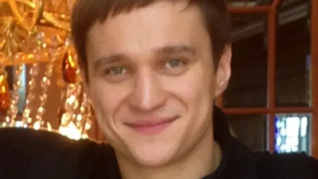 
Um homem de cabelos castanhos curtos sorri em frente a um grande lustre.
