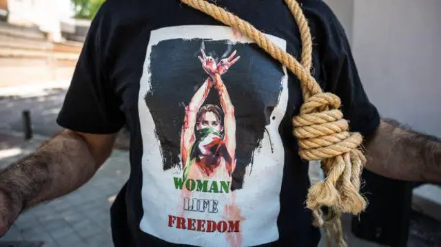 تصویر نمادین فردی که به گردنش طناب دار را آویزان کرده است. این مرد که تنها قفس سینه‌اس دیده می‌شود پیراهنی با نماد «زن، زندگی، آزادی» به تن دارد. 