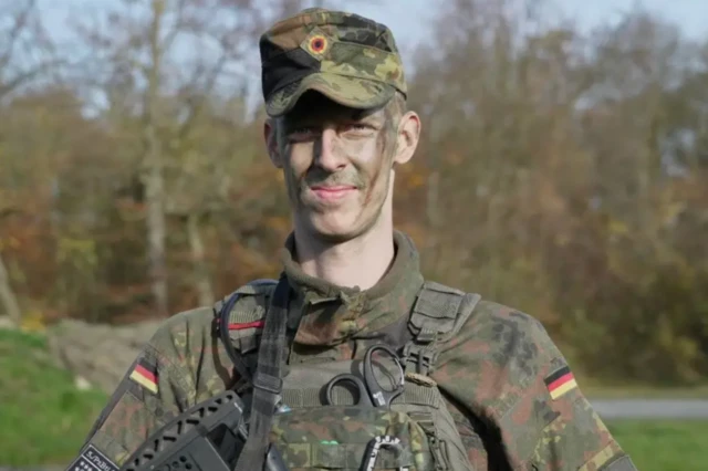 Yüzüne kamuflaj boyaları sürmüş bir Alman askeri BBC haberi için poz veriyor.