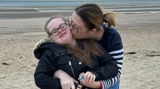 Emily McAllister, wearing a navy jumper, hugging and kissing her daughter Keira, who has down syndrome and is wearing a black coat, as they stand on a sandy beach.