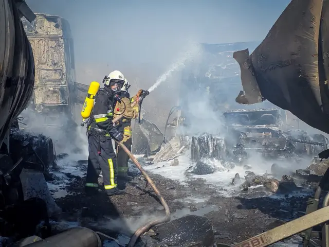 Рятувальники у захисному спорядженні гасять пожежу на зруйнованому промисловому об’єкті, використовуючи шланг із водою або піною.
Довкола видно обгорілі залишки техніки й конструкцій, густий дим і вкриту попелом територію