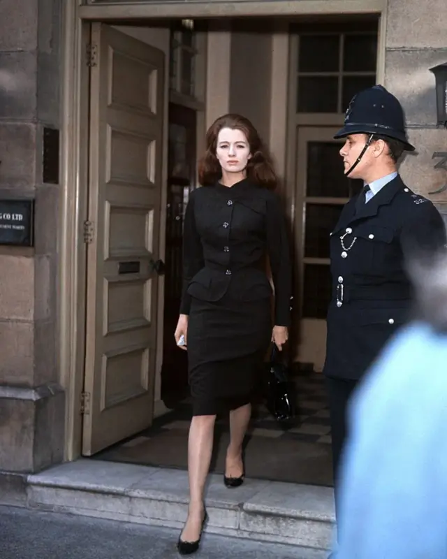 کریسیتن کیلر در حال خروج از یک ساختمان، یک مامور پلیس در کنار در ایستاده است