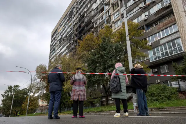 Три женщины и мужчина стоят за ограждением у повреждённого многоэтажного жилого дома. В центре здания видны выбитые окна и следы пожара