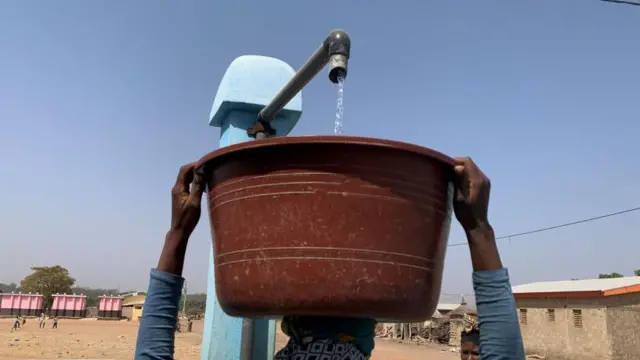 Une femme porte un seau sur la tête, qui est en train d'être rempli d'eau.