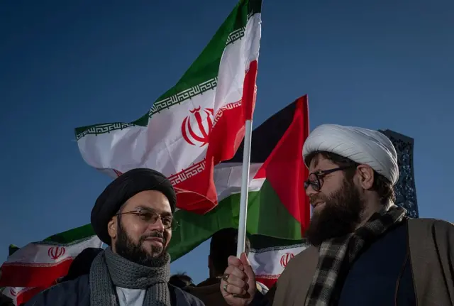 दक्षिणी इरानमा सरकारको पक्षमा निकालिएको एउटा र्‍यालीमा सहभागी दुई धर्मगुरुहरू कुराकानी गर्दै, उनीहरूमध्ये एक जनाले इरानको र अर्काले प्यालेस्टाइनको झण्डा बोकेका छन्