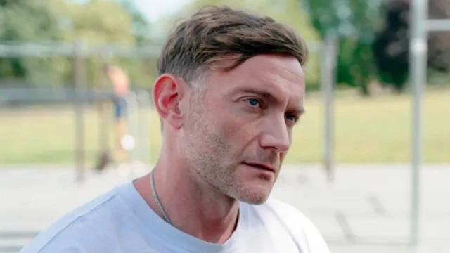 Крупным планом житель Лондона Кристиан Д`Ипполито крупным планом на фоне спортивной площадки. Он смотрит немного сбоку, на лице щетина, он в белой футболке, на шее цепочка серебрянного цвета