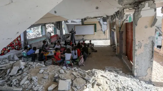 Ғазо Хон Юнисидаги Табария мактаби биносида ерда ўтирган ўқувчилар ва дарс ўтаётган ўқитувчи аёл