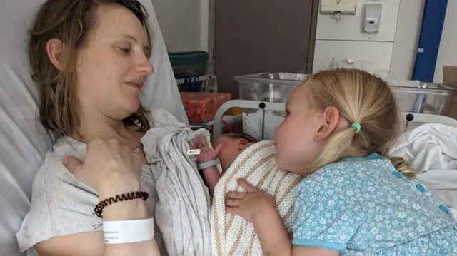Louise en una cama de hospital con su bebé recién nacido y su hija mayor, que lleva un vestido floral azul y tiene cabello rubio.