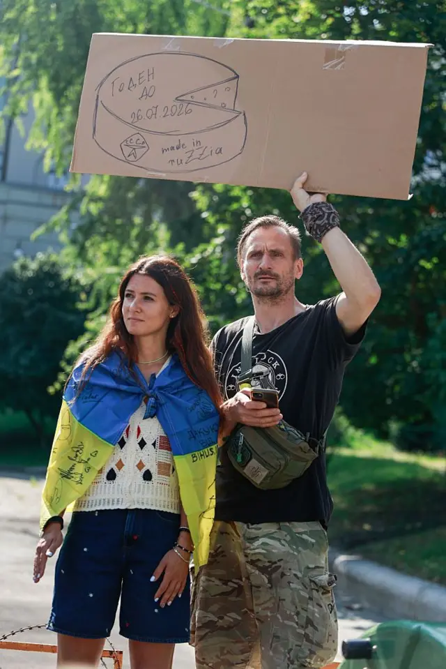Жінка та чоловік на протесті. Чоловік у мілітарі-штанях тримає над головою великий картонний плакат із намальованим кругом сиру та написами, зокрема "made in ruzzia" і "годен до 26.07.2026". Жінка загорнута в український прапор із численними підписами.