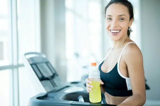 Una mujer en ropa deportiva sonríe mientras sostiene una botella de plástico con una bebida energética de color amarillo fluorescente.