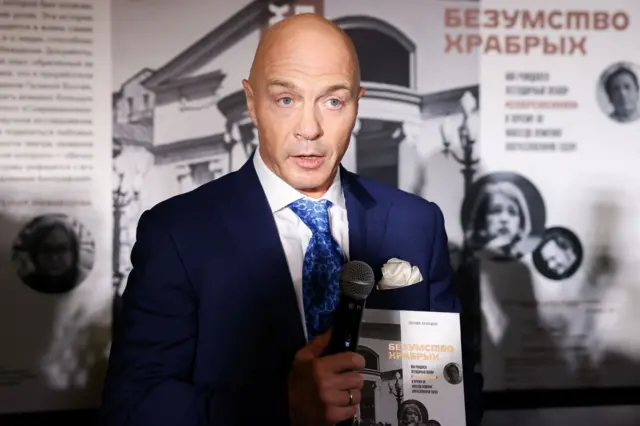 Мужчина в синем костюме держит в руках микрофон и книгу под названием «Безумство храбрых»