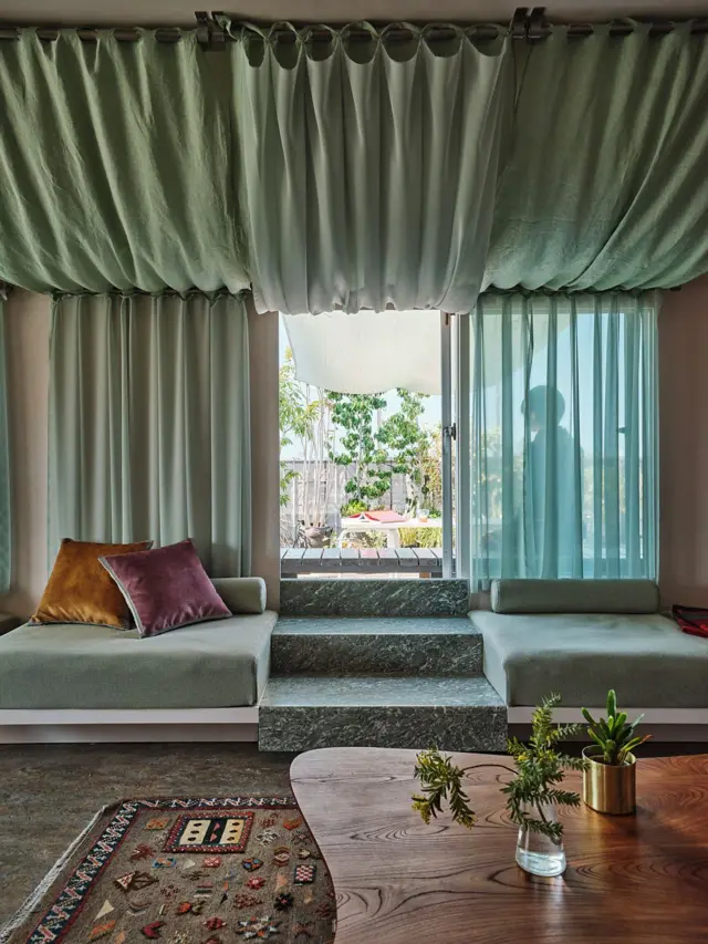 Sala de estar contemporánea con asientos junto a una ventana, cortinas onduladas en el techo, escalones de piedra y mesa de madera.