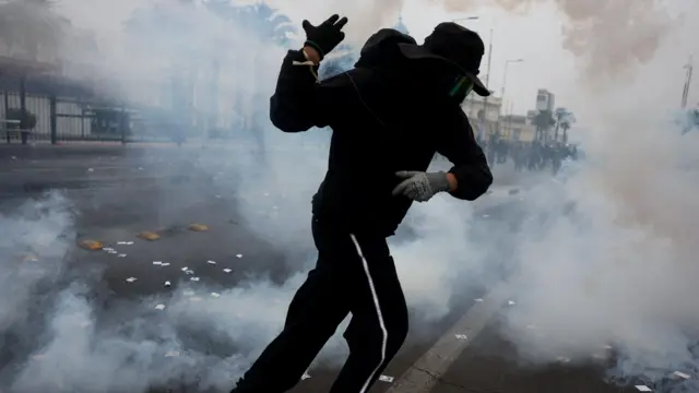 muškarac u crnoj dukserici i trenerci trči kroz oblak suzavca, nosi masku i šešir preko lica, na rukama rukavice