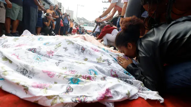 Una mujer besa la cabeza de un ser querido que yace muerto en el suelo y que está cubierto con una tela.