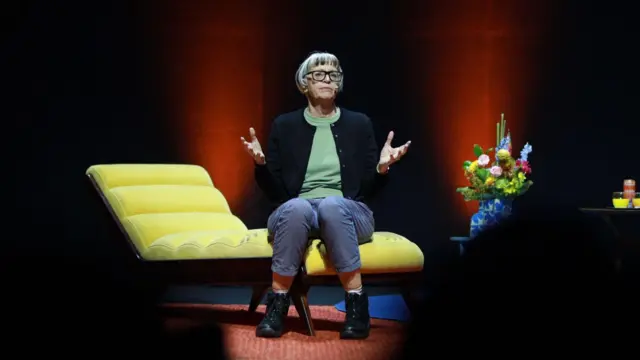 A foto mostra Philippa Perry, uma mulherunibet mmeia-idade, pele clara, cabelos curtos e franja, sentadaunibet mum divã amarelo gesticulando. 