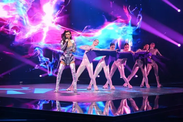 Ива у сребрном костиму, иза ње су плесачице обучене у бела трико одела