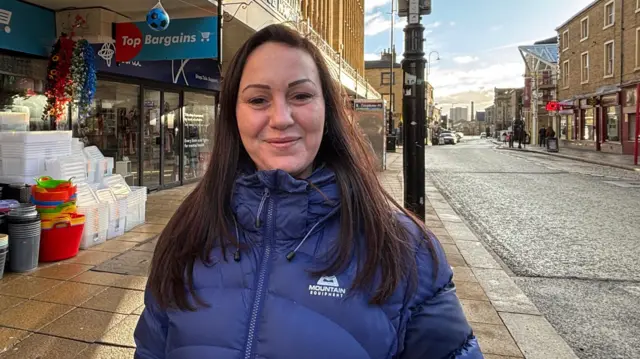 Una mujer que viste una chaqueta acolchada azul y tiene el pelo largo y castaño está de pie junto a una hilera de tiendas que venden cajas y cubos de plástico.