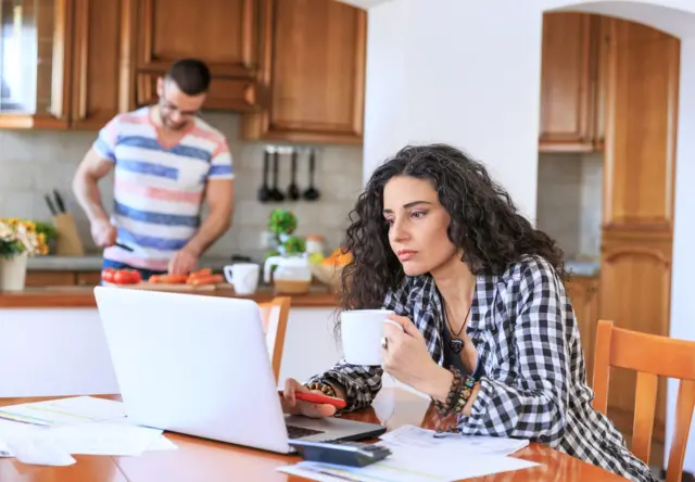 سيدة تستخدم لاب توب ورجل في الخلفية، كلاهما يجلسان في المطبخ يقوم الرجل بتقطيع الخضار يرتدي قميصاً مخططاً بالأبيض والأزرق، بينما تشرب السيدة التي ترتدي قميصاً بلونين أبيض وأسود، القهوة في كوب أبيض وقلماً بلون أحمر أثناء عملها.
