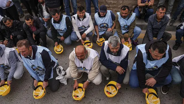Maden işçileri ellerindeki sarı renkli baretleri yere vuruyor. Üzerlerinde beyaz renkli yelekler var.