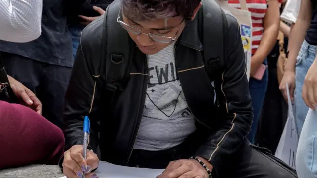 Una persona firma un documento. Está en primer plano, tiene anteojos.